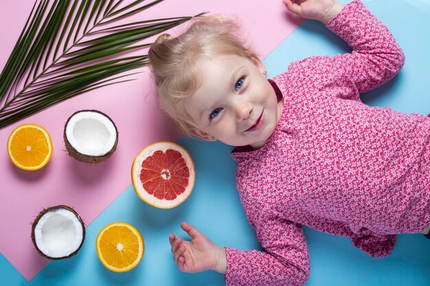 Petite fille souriante allongée près de fruits exotiques sur fond bleu. vue de dessus, mise à plat.