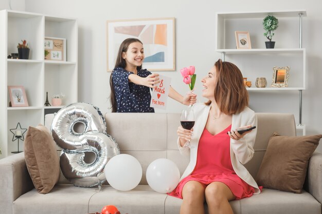 Une petite fille souriante debout derrière un canapé donne des frondes avec une carte postale à une maman souriante avec un téléphone et un verre de vin dans le salon