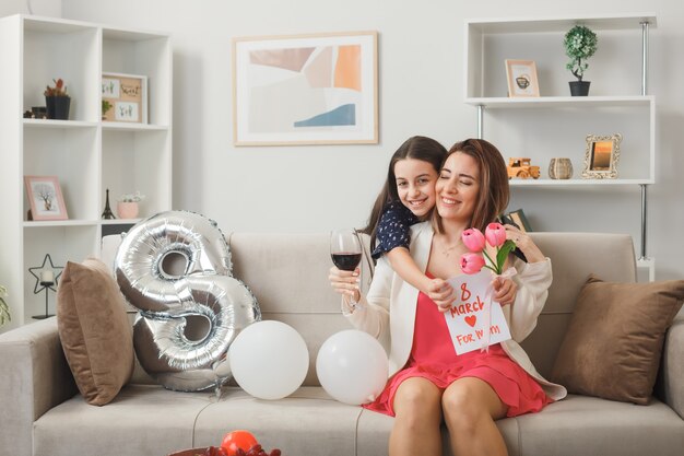 Une petite fille souriante debout derrière un canapé tenant des fleurs avec une carte de voeux a embrassé sa mère sur un canapé le jour de la femme heureuse dans le salon