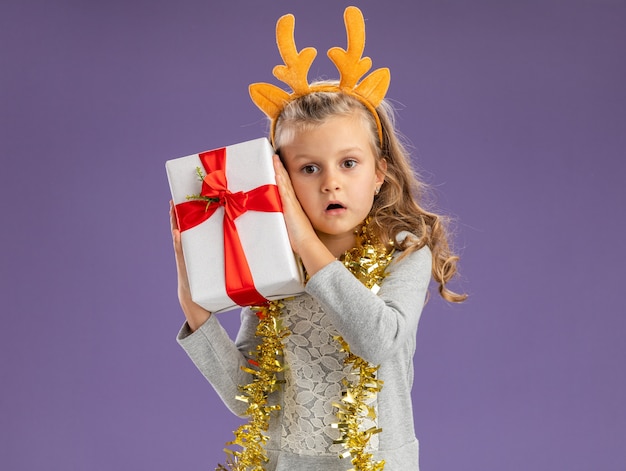 petite fille surprise portant un cerceau de cheveux de noël avec une guirlande sur le cou tenant une boîte-cadeau autour du visage isolé sur un mur bleu