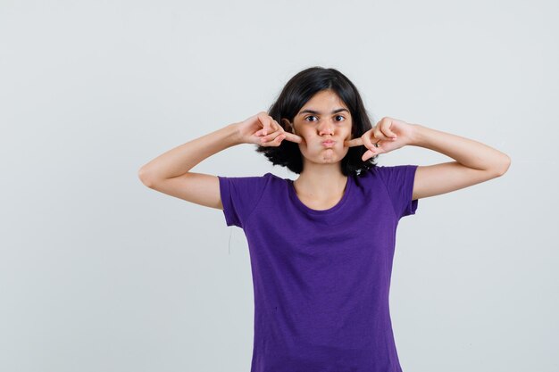 Petite fille en t-shirt en appuyant sur les doigts sur les joues soufflées,