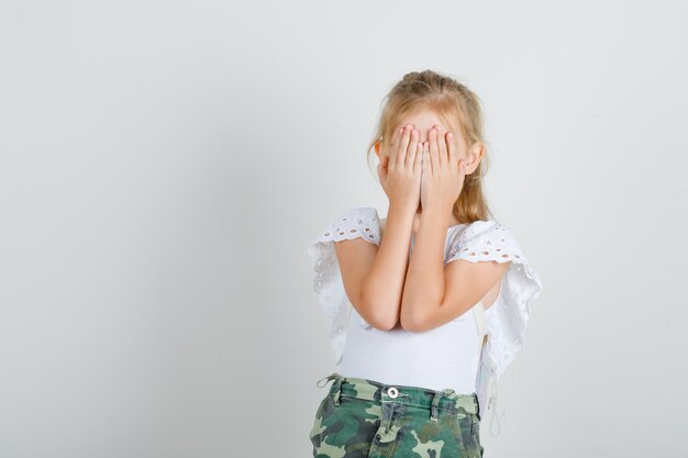 Petite fille en t-shirt blanc, jupe couvrant les yeux avec les mains