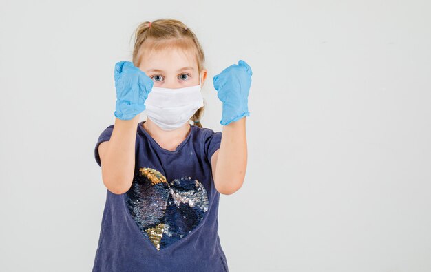 Petite fille en t-shirt, gants et masque levant les mains avec le poing fermé et à la vue de face déterminée.