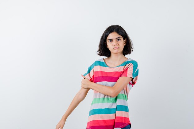 Petite fille en t-shirt gardant la main sur l'autre bras et l'air sérieux, vue de face.