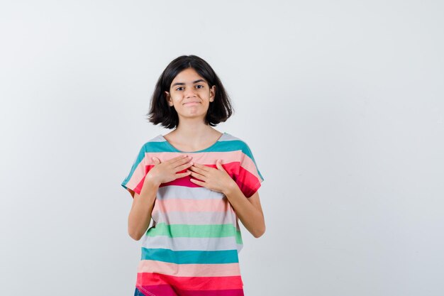 Petite fille en t-shirt gardant les mains sur la poitrine et regardant joyeusement, vue de face.