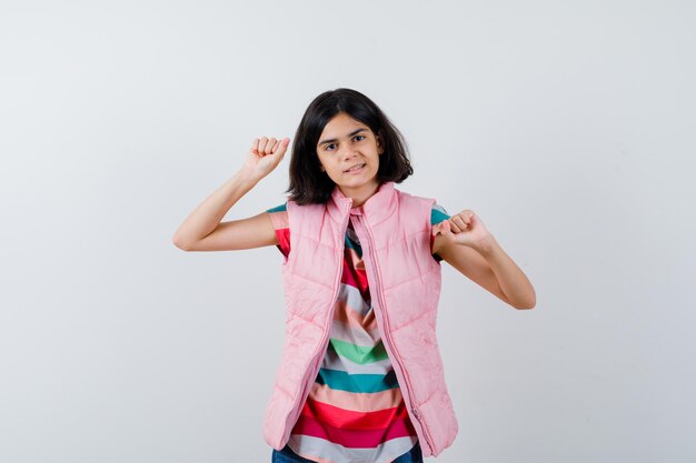 Petite fille en t-shirt, gilet matelassé, jeans montrant le geste du gagnant et l'air heureux, vue de face.