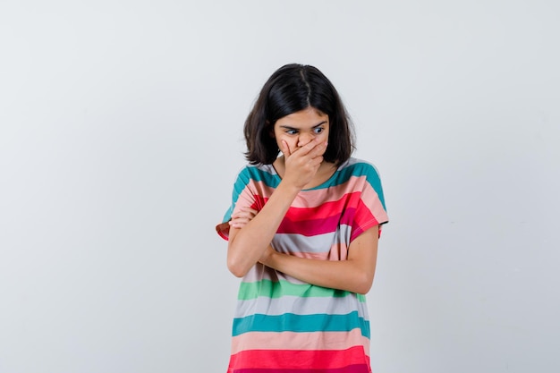 Petite fille en t-shirt, jeans couvrant la bouche avec la main tout en tenant une main sur l'avant-bras et l'air surpris, vue de face.