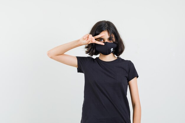 Petite fille en t-shirt noir, masque montrant le signe v près de l'œil et l'air confiant, vue de face.