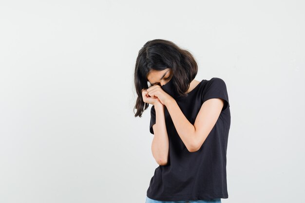 Petite fille en t-shirt noir, masque penchant le visage sur les mains jointes et à la vue épuisée, de face.