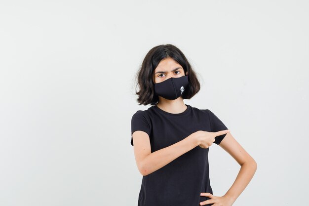 Petite fille en t-shirt noir, masque pointant vers le côté et regardant sérieusement, vue de face.