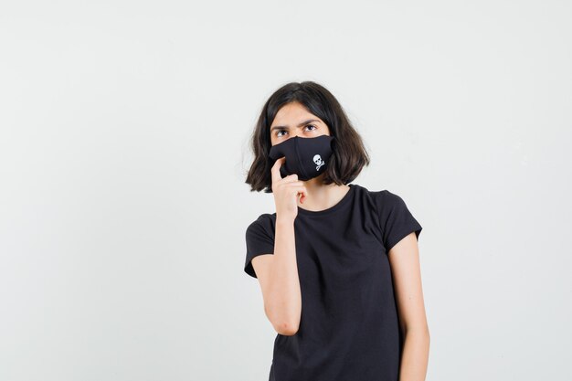 Petite fille en t-shirt noir, masque à la recherche et à la vue réfléchie, de face.