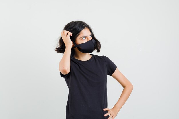 Petite fille en t-shirt noir, masque se gratter la tête et regardant pensif, vue de face.