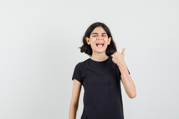 Petite fille en t-shirt noir montrant le pouce vers le haut et regardant heureux, vue de face.