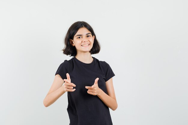 Petite fille en t-shirt noir pointant vers l'avant et regardant heureux, vue de face.