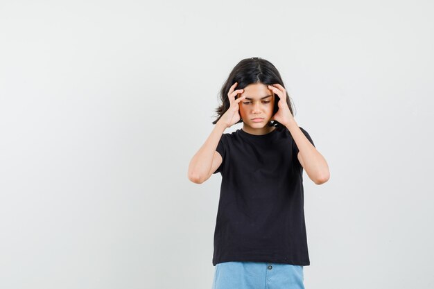 Petite fille en t-shirt noir, short ayant de forts maux de tête et regardant malade, vue de face.