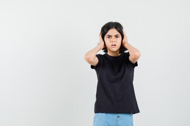 Petite fille en t-shirt noir, short tenant les mains sur les oreilles et à la peur, vue de face.