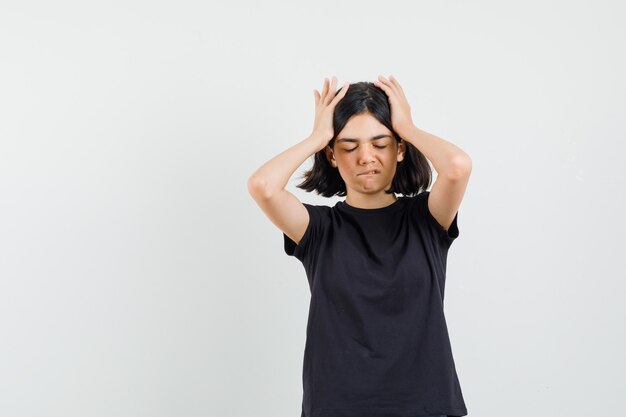 Petite fille en t-shirt noir, tenant les mains sur la tête, mordant la lèvre et regardant oublieux, vue de face.