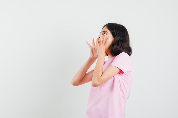 Petite fille en t-shirt rose criant avec les mains près de la bouche.