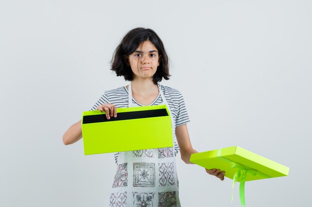 Petite fille en t-shirt, tablier tenant une boîte cadeau ouverte et à la confusion,