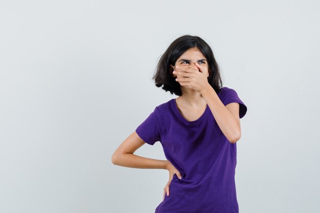 Petite fille en t-shirt tenant la main sur la bouche et à la joyeuse,