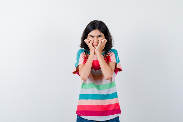 Petite fille en t-shirt tenant les mains sur les joues et l'air heureux, vue de face.