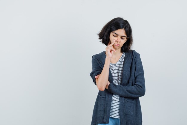 Petite fille en t-shirt, veste en appuyant sur le doigt sur la joue et à la tristesse,