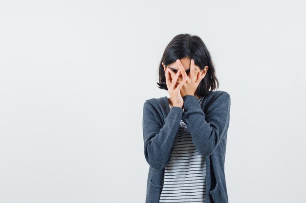 Petite fille en t-shirt, veste regardant à travers les doigts et à la curiosité