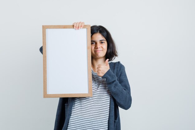 Petite fille en t-shirt, veste tenant un cadre vide, montrant le pouce vers le haut et à la joie,