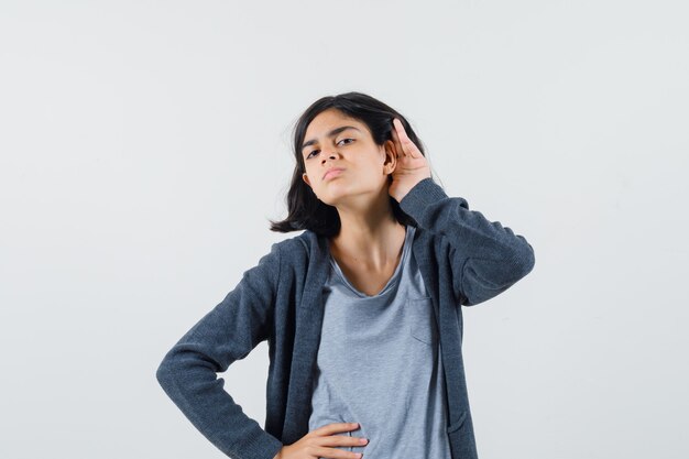 Petite fille en t-shirt, veste tenant la main derrière l'oreille et à la curiosité