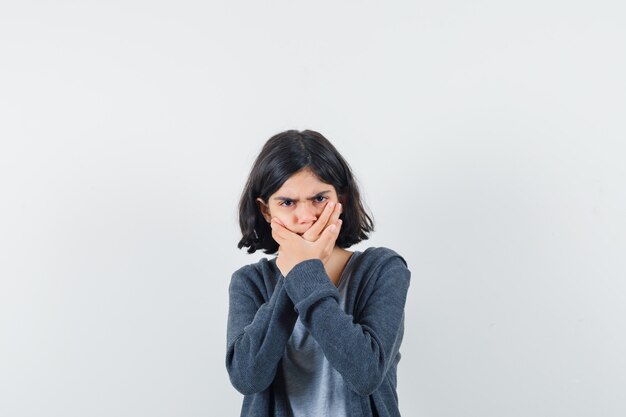 Petite fille en t-shirt, veste tenant les mains sur la bouche et à la recherche d'anxiété