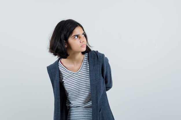 Petite fille en t-shirt, veste tenant les mains derrière le dos et à la recherche concentrée,