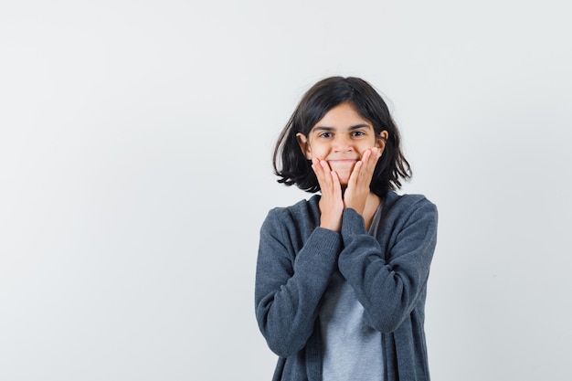 Petite fille en t-shirt, veste tenant les mains sur les joues et à la joyeuse vue de face.