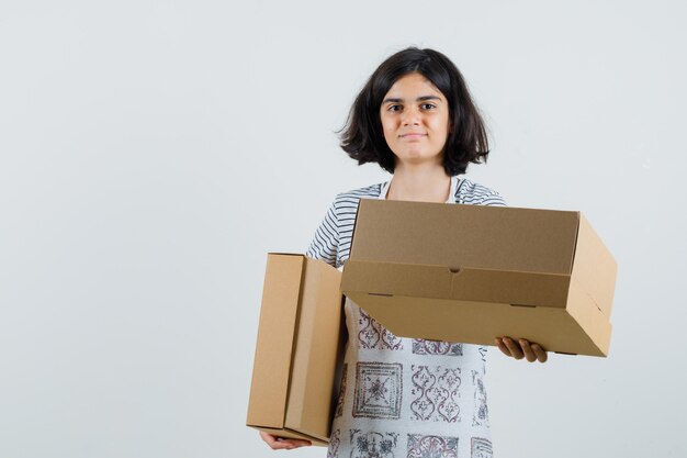 Petite fille tenant des boîtes en carton en t-shirt, tablier et à la joyeuse.