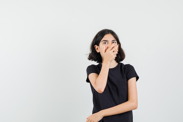 Petite fille tenant la main sur la bouche en t-shirt noir et à la vue étonnée, de face.