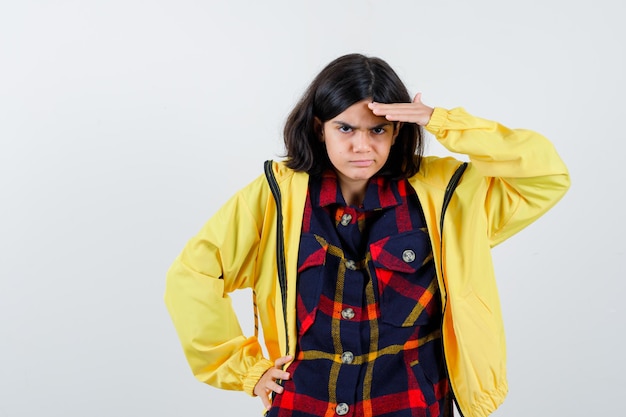 Petite fille tenant la main sur le front à la recherche de manière en chemise à carreaux, veste et l'air concentré, vue de face.