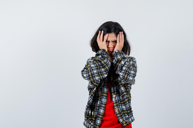 Petite fille tenant la main sur sa tête en chemise, veste et semblant anxieuse, vue de face.