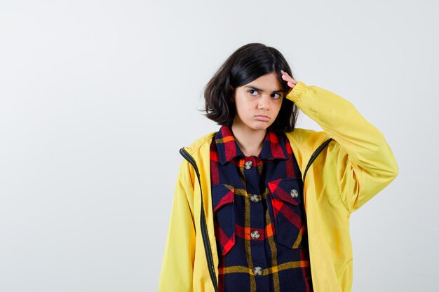 Petite fille tenant la main sur la tête en chemise à carreaux, veste et à l'air indécise. vue de face.