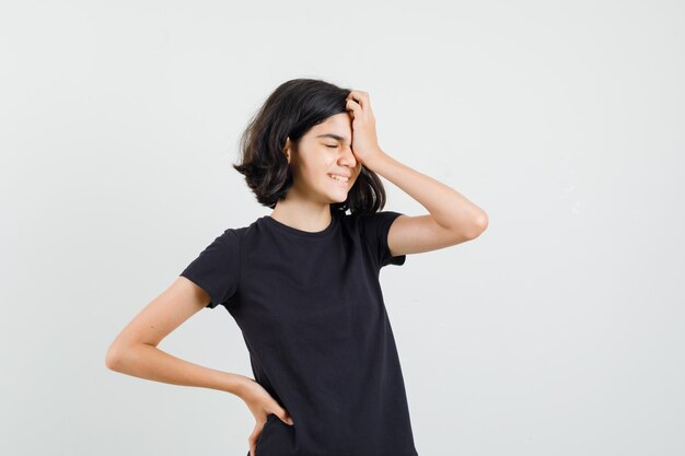 Petite fille tenant la main sur le visage en t-shirt noir et à la honte, vue de face.