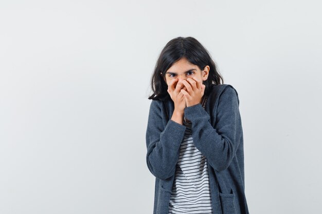 Petite fille tenant les mains sur la bouche en t-shirt, veste et à la recherche excitée.