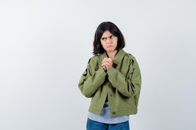 Petite fille tenant les mains jointes sur la poitrine en manteau, t-shirt, jeans et hésitante. vue de face.