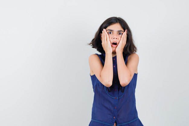 Petite fille tenant les mains sur les joues en chemisier bleu et à la recherche alarmée. vue de face.