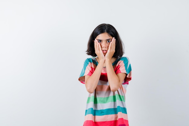 Petite fille tenant les mains sur les joues en t-shirt, jeans et l'air surpris. vue de face.