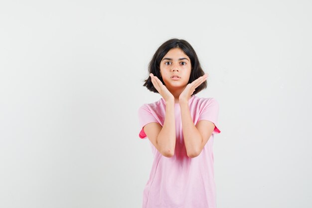 Petite fille tenant les mains près du visage en t-shirt rose et à la perplexité. vue de face.