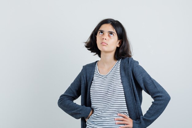 Petite fille tenant les mains sur la taille en t-shirt, veste et à la recherche indécise.