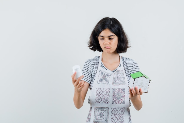 Petite fille tenant le modèle de maison, bouteille de pilules en t-shirt, tablier et à la recherche hésitante.