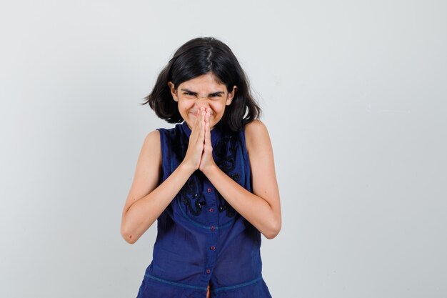 Petite fille tenant par la main dans le geste de prière en chemisier bleu et à la joyeuse vue de face.