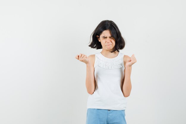 Petite fille tenant les poings serrés en chemisier blanc, short et à la méchante, vue de face.