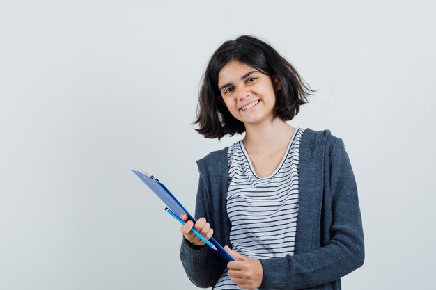 Petite fille tenant le presse-papiers avec un crayon en t-shirt, veste et à la joyeuse,