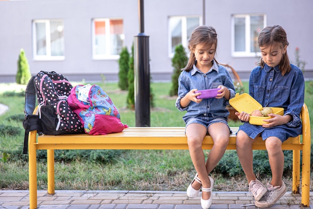 Petites écolières assises sur un banc dans la cour de l'école et mangeant dans des boîtes à lunch.