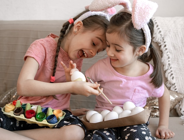 Petites sœurs en oreilles de lapin peignent des oeufs de Pâques sur le canapé à la maison